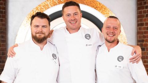 The three finalists from left to right, Mark O'Brien, Gareth Baty and Luke Emmess. All three chefs are wearing white chefs' coats and black trousers. O'Brien has short dark hair and a full beard. Baty, who has his arms round the shoulders of the other two, is taller and has similar short dark hair with light stubble. Emmess has very short dark hair on the top of his head and is shaved at the sides. He has a short goatee beard. They are on the set of MasterChef, with a brick wall and the programme's logos behind them.