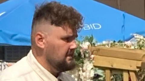 Albert Jarosz: A profile view of a man with dark hair, shaved at the side, and dark beard. Behind him is a blue umbrella with the letters "BID" written on it in read.
