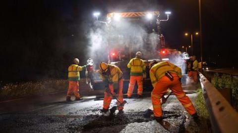 Workers resurfacing a road at night. Two are spreading tarmac with brushes while other workers gather around a large vehicle, which has bright lights near the top.