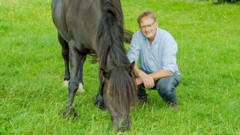 Roger Sewill smiling for a photo as he crouches in a field next to Scarlet, a brown Dales pony, which is grazing on green grass.
