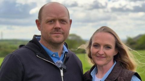 Dicky and Clare Lanham stand in a green field. Dicky is on the left of frame, wearing a navy blue zipped jacket on top of a light blue polo shirt. Clare is on the right of frame. She wears a gilet over a light blue shirt.