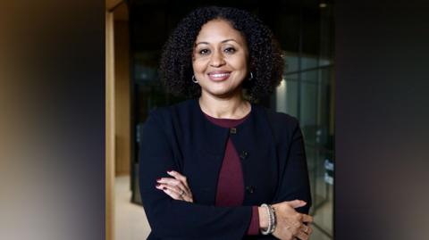 Andrea Simon in a dark suit smiles at camera with arms crossed 