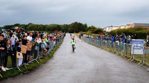 A child riding a bicycle on a pathway with crowds of people supporting from the side lines. 