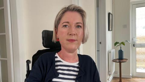 Photo of Sarah, who is looking into the camera and smiling slightly. She has short, chin-length light blonde hair, and is wearing a pair of red earrings. Sarah is wearing a navy blue and white striped top, with a dark blue cardigan over the top. She is pictured in her powered wheelchair, with the back of the chair and the headrest just visible over her shoulder. The photo has been taken indoors, inside her home, which has white walls and a brown wooden floor. On the right of the photo, a glass-panelled door is seen, and next to it is a brown wooden table with a green leafy pot plant on it in a blue vase. 