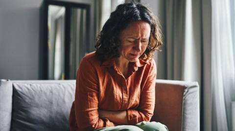Stock image of woman with curly brown shoulder length hair sitting on a grey softa. Her arms are folded over her abdomen and she appears to be grimacing and upset in pain. She wears a terracotta shirt and green trousers. There is light coming in through a nearby window. 