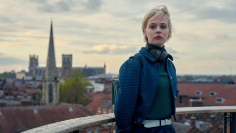 A woman with headphones around her neck stands on a balcony overlooking York city centre and York Minster.