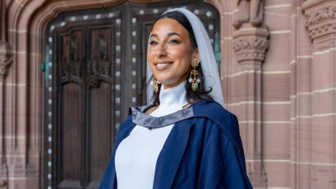 Bride and graduate Charnele Washbourne wearing a wedding dress and veil with her graduation gown and gold and green dangle earrings inside Liverpool Anglican Cathedral. She is looking away from the camera. She is smiling.