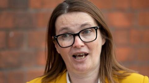 A woman talking. Former politician Kirsty Williams has black framed glasses on and short light-brown hair. She has a yellow jacket on, under which is a white shirt.