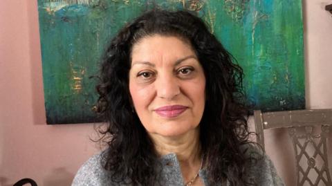 Nashmin Riazi pictured looking straight into the camera. She has curly shoulder length black hair, wears pink lipstick, a grey cardigan and white shirt underneath.