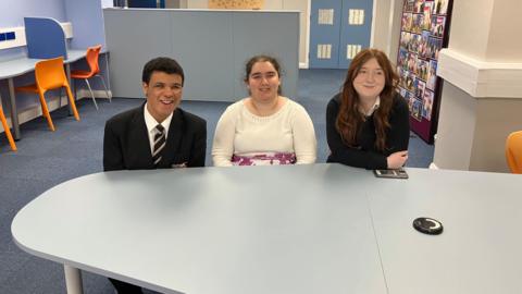 Three teenagers sit at a large desk in a classroom. On the left, a boy with short dark hair is wearing a dark suit with a white shirt and a striped tie, a girl in the middle with tied-back dark curly hair is wearing a cream jumper and on the right, a girl with long dark red hair is wearing a white blouse under a black jumper. 