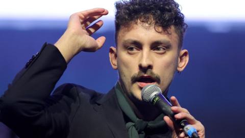 Antony Szmierek, with curly short hair, wears a black jacket and green scarf and speaks into a microphone