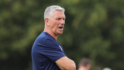 John Askey shouts orders on training ground with arms crossed in blue club training t-shirt