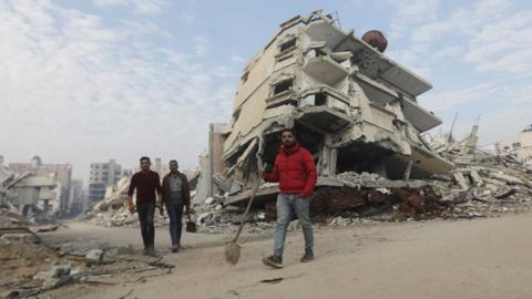 A man in a read puffer jacket stands in a ruined street in Gaza. He is holding a shovel.