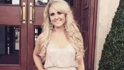 A slightly blurry photo of Amber Rose Cliff who has long blonde curly hair and is smiling into the camera. She is wearing large, silver earrings and a light pink dress which is cinched at the waist. Her left hand is on her hip. She is standing in front of dark, wooden doors and there is a hedge to the right.