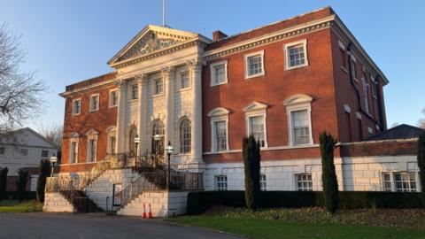 A view of Warrington Town Hall. It is a Georgian building with a large staircase leading up to the front. The centre of the building at the front has four white pillars.