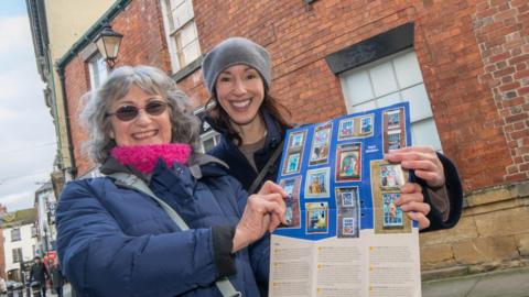 Councillor Hannah Gostlow and Liz Baxandall hold up a window trail programme, outside a building which features one of the artworks.