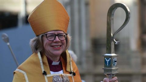 Dame Sarah Mullally wears gold ceremonial garments and a mitre, holding a curved staff, with stone buildings visible in the background, as she departs after her installation as the Archbishop of Canterbury.