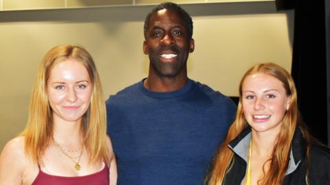 Dwain Chambers in the middle of the picture wearing a navy jumper. Either side of him are two young women. The one on the left of the image is wearing a red vest while the one on the right is wearing a black coat. They are all smiling and looking into the camera.