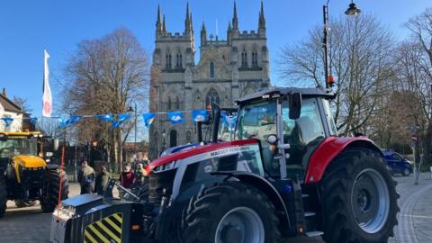 Two tractors parked outside a church in a town. In the background there are people and a string of blue flags hanging in the air. 