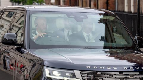 Prince Harry can be seen in the backseat of a Range Rover as it drives outside Clarence House, with two figures in the front seats, pictured through the windscreen with the glass reflecting the sky.