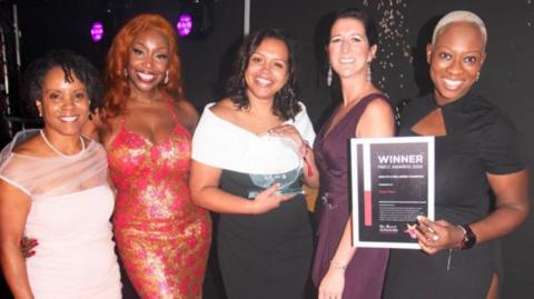 Five women posing against a black backdrop, with the woman on the right hand side holding up a paper that says winner. The women are dressed in ballgowns 