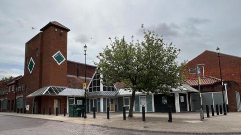 The City Square shopping centre, which has been closed since 2019. It is a red-brick building with a three-storey tower and angular window features. Outside is a tiled square, dominated by a tree with green leaves.