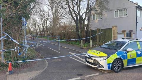 A police car is on the right of the picture, sitting in front of a road that has blue and white police tape across it. The small road has trees either side, and houses are behind the fences.