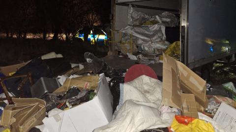 A large number of waste, lying on the ground, showing boxes, a chair, bedding, in front of a white van, with its back open. A police car is in the distance but trees and bushes.