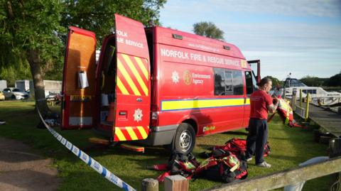 An emergency services worker stands on the grass near a red Norfolk Fire and Rescue Service van. A boat can be seen in the background. Police tape can be seen in the foreground.