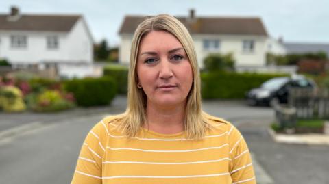 Lisa Blondel - a woman with blonde shoulder length hair, looking sternly towards the camera. She is wearing a yellow and white striped jumper. 