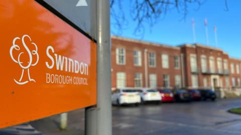 An orange sign has Swindon Borough Council written on it. In the background is a red bricked council office with cars parked outside it on a road.