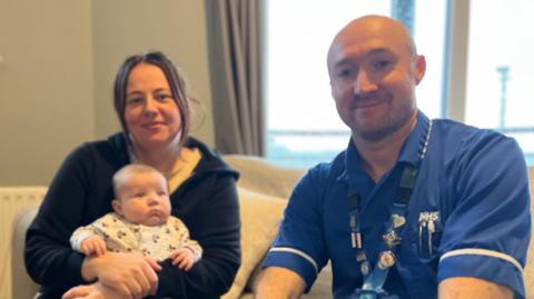 12-week old Myles is wearing a white baby grow and is being held by his mother. His mother, Shanice, is wearing a black zip hoodie and has brunette hair. Sat next to her is Chris in blue nurses scubs. He is smiling and has very short hair.