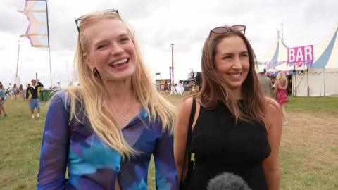 A wide shot of Emma and Kate. Emma is blonde with glasses on her head, and is wearing a blue top. Kate has long brown hair and is wearing a dark top. In the background are various tents.
