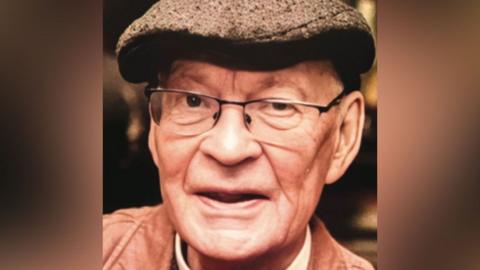 Close up photo of Right Reverend Stephen Pedley. He's wearing a brown flat cap, glasses a brown jacket and red jumper with a clerical dog collar around his neck.