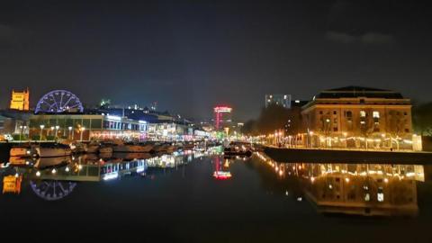 A nighttime image of Bristol harbour - with a big expanse of water, which is in dark, and lit up buildings in the background