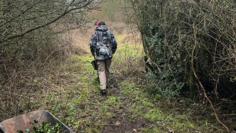A person pictured from the back who is walking with a wheelbarrow in a wooded area. There are among trees and near to the camera is part of another wheelbarrow which has leaves in it. 