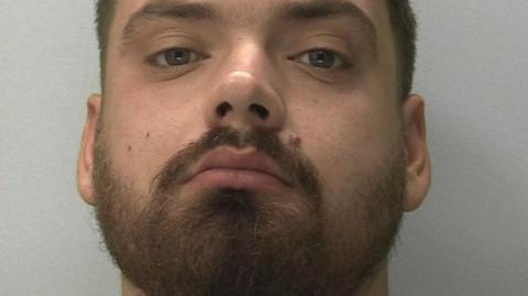 Ben Helliker - aman with a dark beard, moustache and short dark hair is wearing a grey T-shirt. He is looking at the camera with slightly downcast eyes.