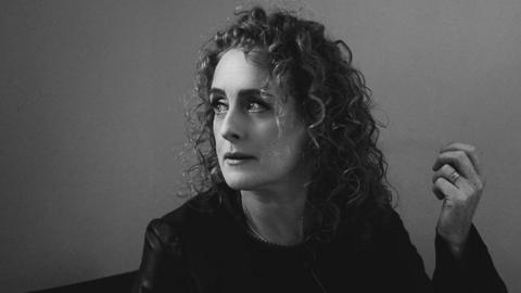 A black and white headshot of a woman - Briana Corrigan - looking to the side. She has curly hair, is wearing a jacket and is holding her left hand up. 