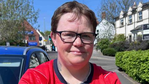 A person wearing a red England football shirt with short hair and glasses is standing on a residential street