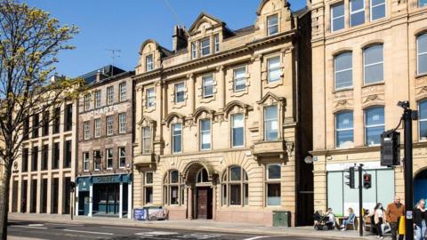 A row of historic-looking buildings along a street. In front of one of the buildings, people sit at outdoor tables. The four-storey buildings are in different styles. Coronation Buildings, in the centre, is made of large, light brown stone with mainly sash windows. On the ground floor there is a large wooden central doorway beneath an arch, with arched windows immediately either side and then rectangular ones at each end. There is ornate masonry and decorative pillars on the two upper floors which each contain five windows. Above that, within the roof space, is a large, three-sectioned window beneath a triangular design in the middle, and smaller single windows under an arched dormer at each end.