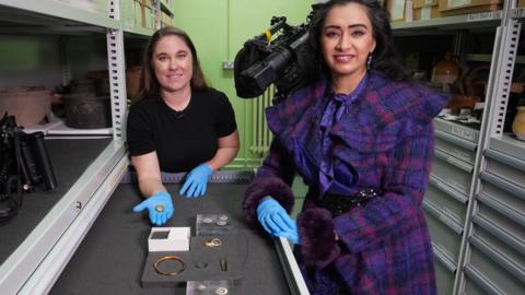 Presenter Roo Irvine with Herefordshire Council’s Museums Service Collection and Engagement Curator - Archaeology, Dr Katie Miller, and some of the Herefordshire Hoard. Roo is wearing a pink and purple coat, and blue plastic gloves. Miller, in a black t-shirt and blue plastic gloves, holds out one of the gold coins.