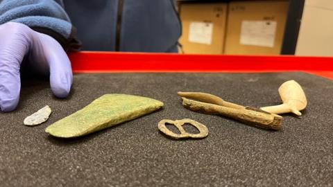 Five objects which have been placed on a grey surface. From far left they show a small grey arrowhead, an axe head, a buckle, nutcrackers and a clay pipe. In the background is a person's hand with a lilac latex glove on. 
