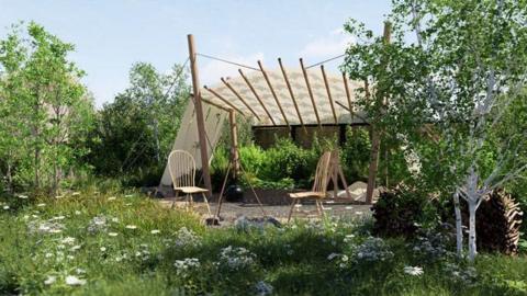 A garden with white flowers among long grass, trees on either side and a canopy structure shading two wooden chairs in the middle.