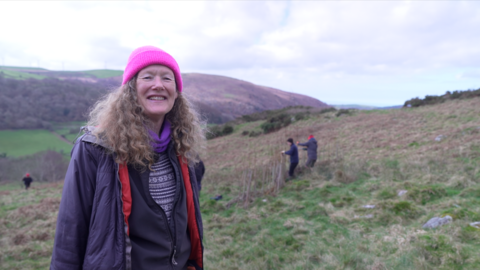 Woman with long, light brown, curly hair is standing on a hillside. She's wearing a bright pink beanie hat and a dark coloured coat. She's smiling at the camera. In the background you see three people on the hillside. Two are working on creating a dam from willow stakes.