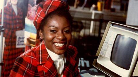 A woman looks into the camera with a big smile. She is wearing a tartan uniform with a tartan beret. 