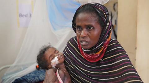Touma, a Sudanese mother wearing a headscarf holding her baby who has a drip in her nose