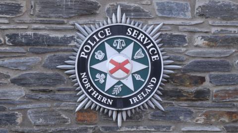 On a grey and brown brick wall is the Police Service of Northern Ireland badge. The badge is circular and it has silver spikes around it, the first section of the circle is black with white writing saying 'Police Service Northern Ireland'. The next circle inside this is green with six symbols on it in white. Then there is six white spikes with another circle inside which has a red cross.