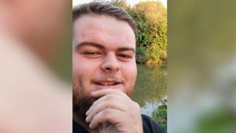 A selfie of Liam Rowe standing beside a lake and some trees, with his hand resting on his chin and smiling at the camera.