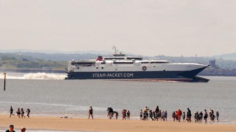 A ferry jets off while in the foreground people are stood on the beach watching it go by. 