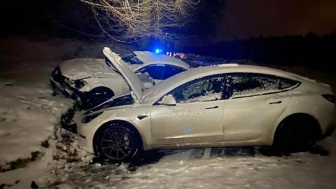 Two white cars are pictured in the snow. They are damaged following a crash. Blue emergency service lights can be seen in the distance.